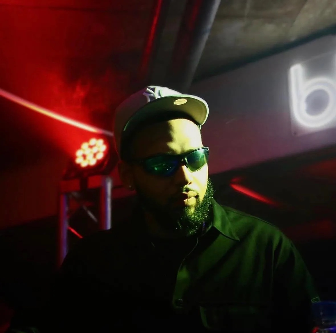 A DJ in a cap and sunglasses lit red, with stage lights and a neon sign behind him