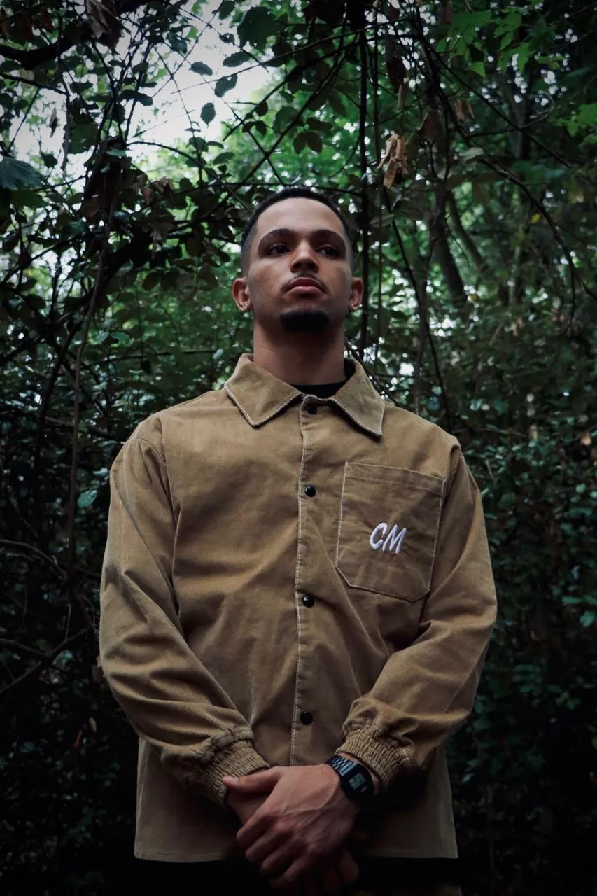 A man in a tan corduroy shirt with a CM monogram, standing in dense green foliage