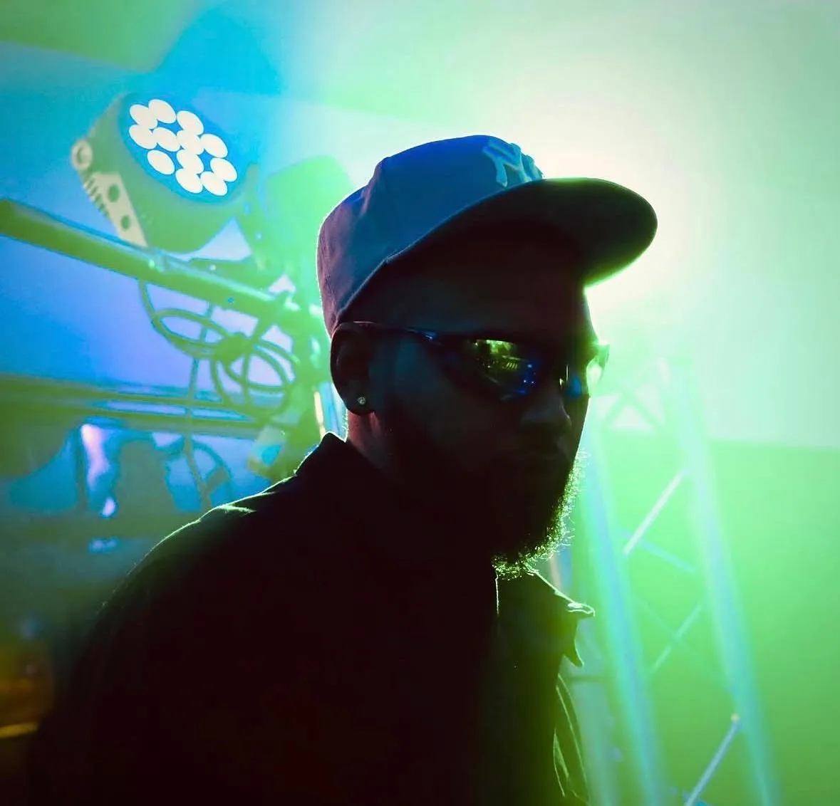 A DJ in a cap and wraparound sunglasses under green and blue stage light