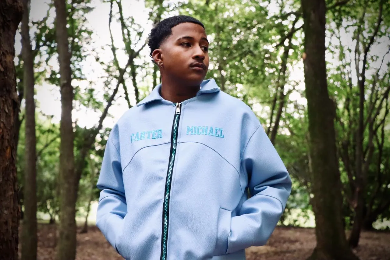 A young man in a pale blue Carter Michael track jacket, hands in pockets, among trees