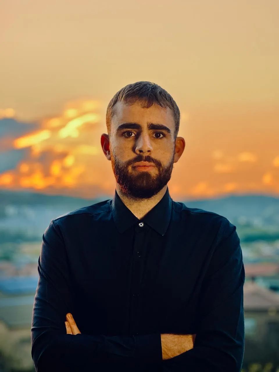 A bearded man in a dark shirt with his arms crossed, in front of an orange sunset
