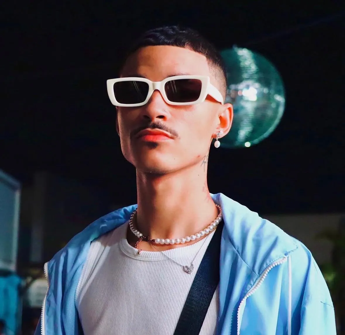 A young man in white sunglasses and a pearl necklace, with a mirror ball behind him
